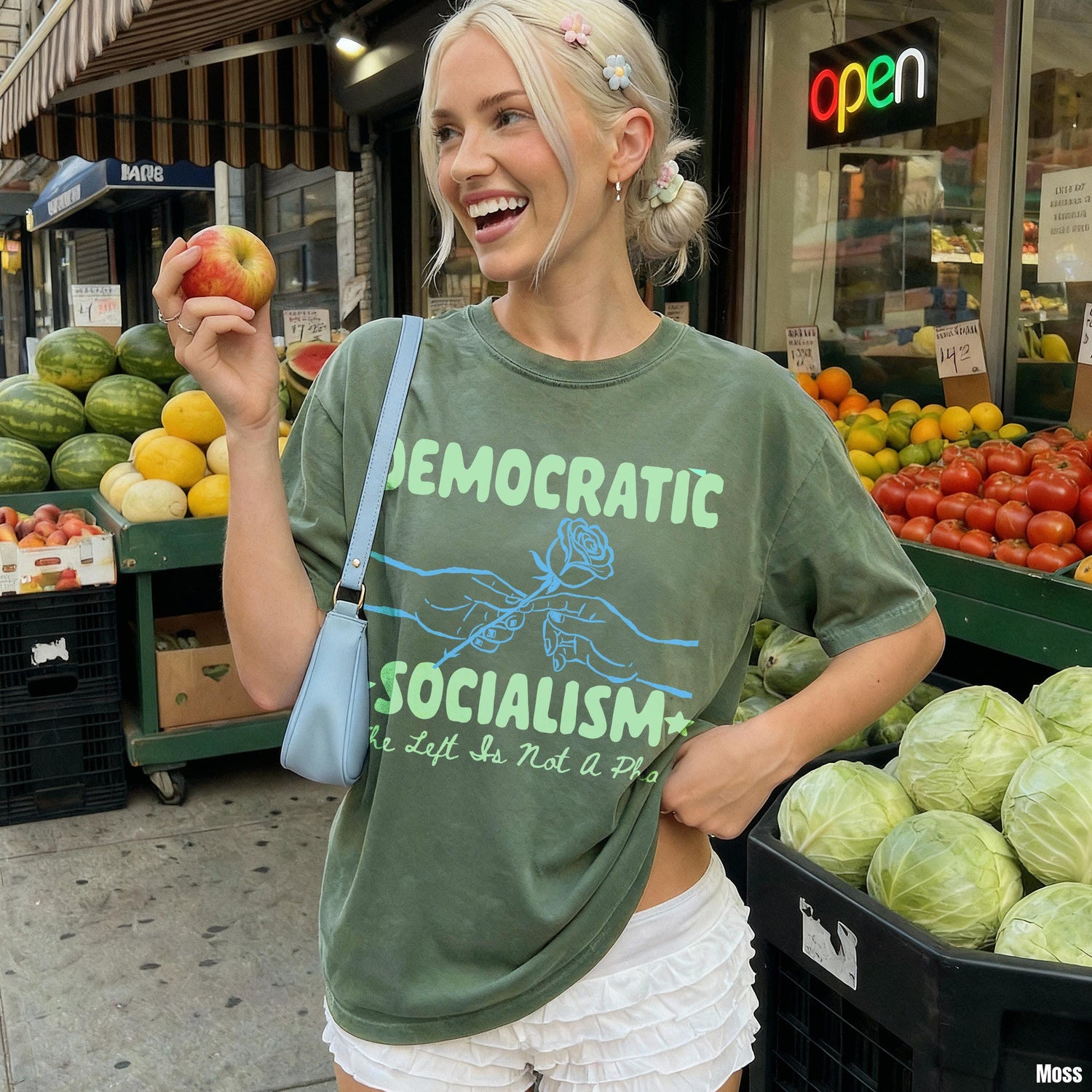 Woman wearing green t-shirt with &amp;quot;Democratic Socialism&amp;quot; text, holding an apple by fruit stand.