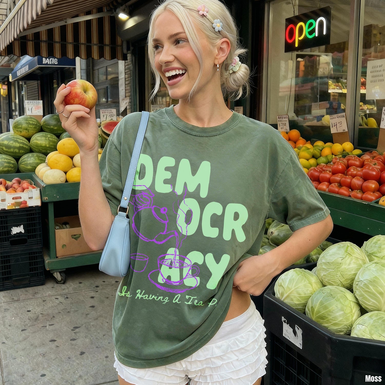 Green t-shirt with &amp;quot;DEMOCRACY&amp;quot; and tea graphic, held by a woman at a fruit stand.