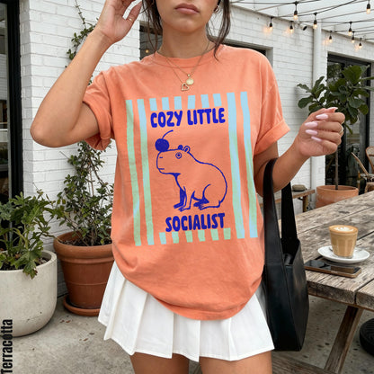 Peach t-shirt with graphic of capybara wearing cherry and text &amp;quot;Cozy Little Socialist.&amp;quot;