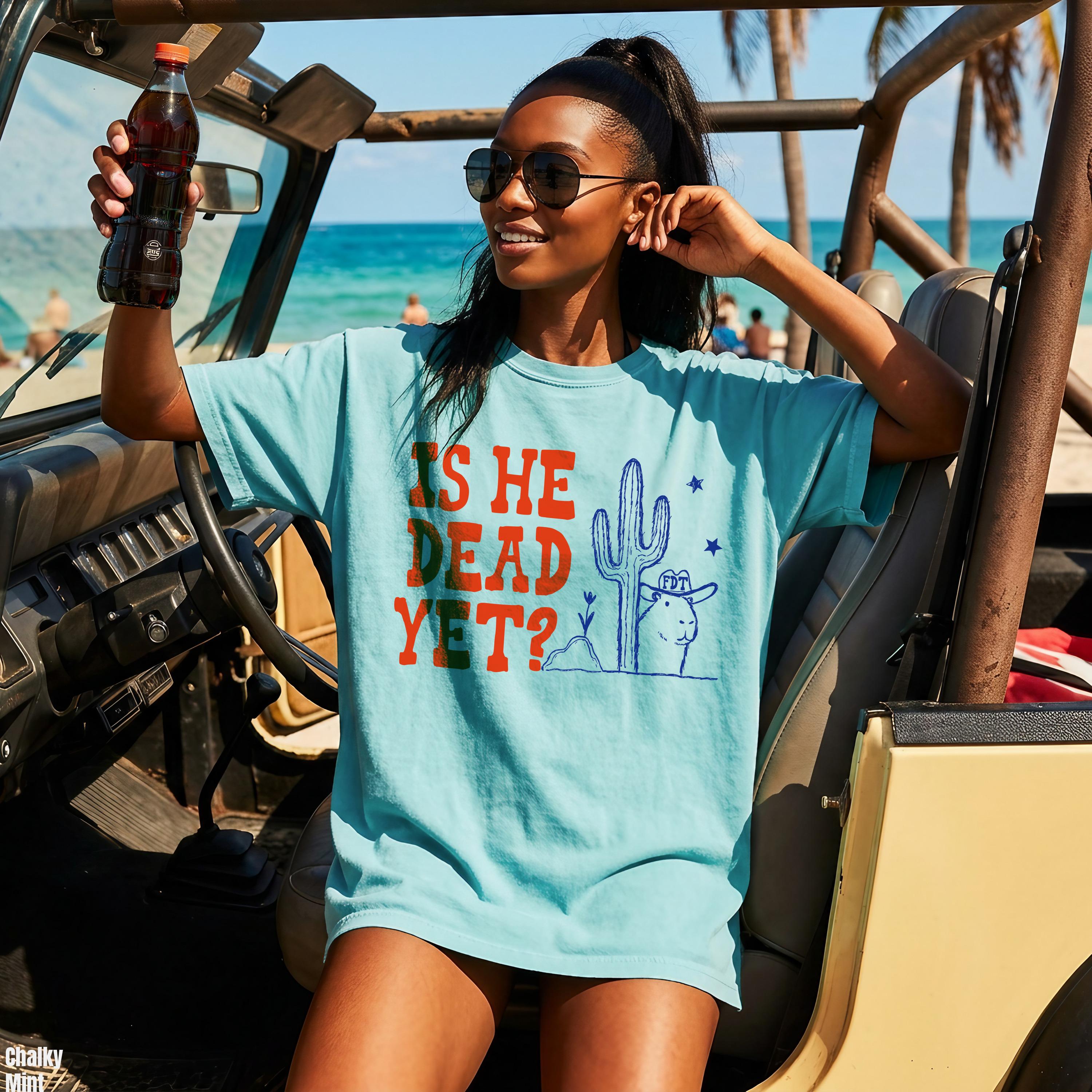 Light blue t-shirt with red text &amp;quot;IS HE DEAD YET?&amp;quot; and desert graphic.