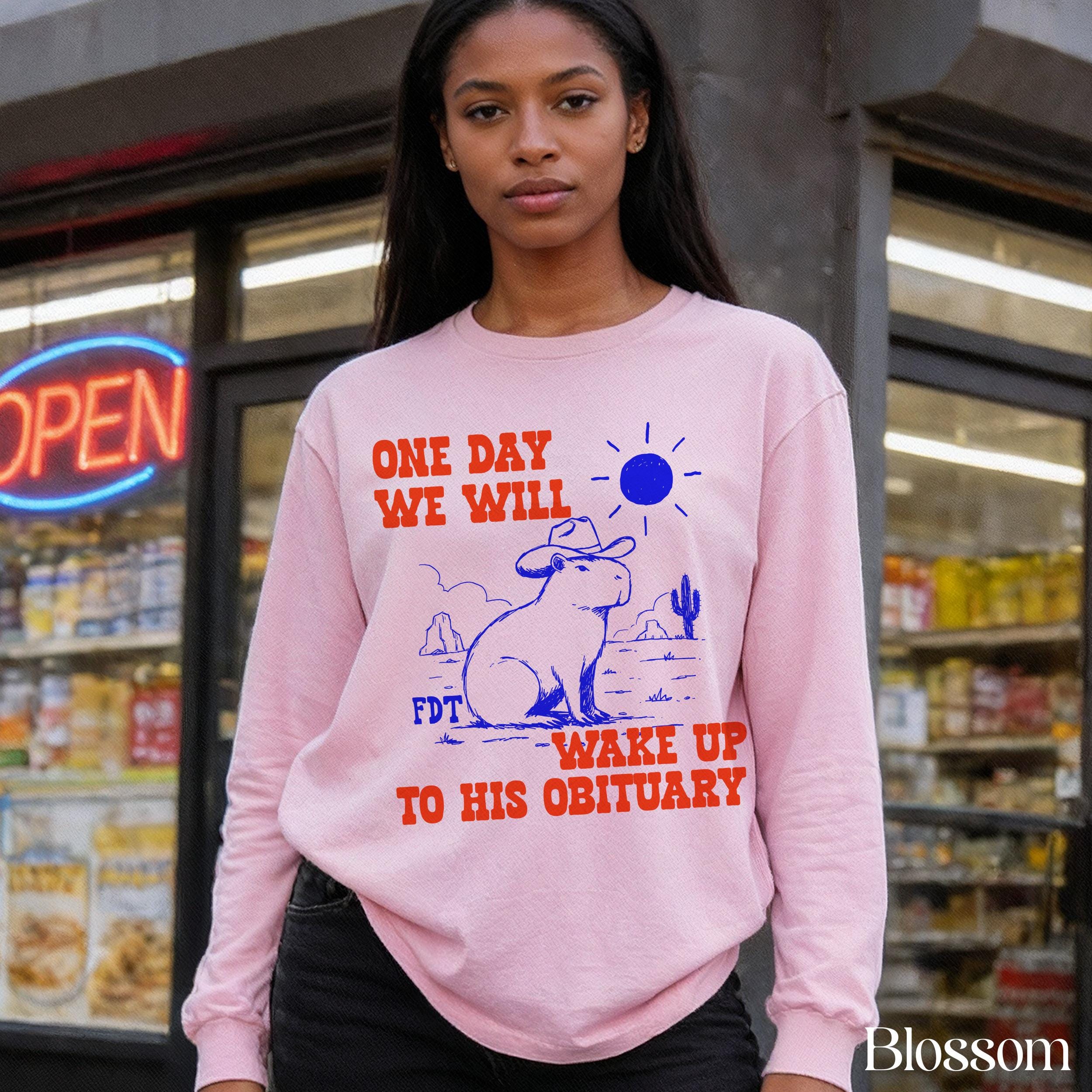 Pink sweatshirt with capybara graphic and text &amp;quot;ONE DAY WE WILL WAKE UP TO HIS OBITUARY&amp;quot;.