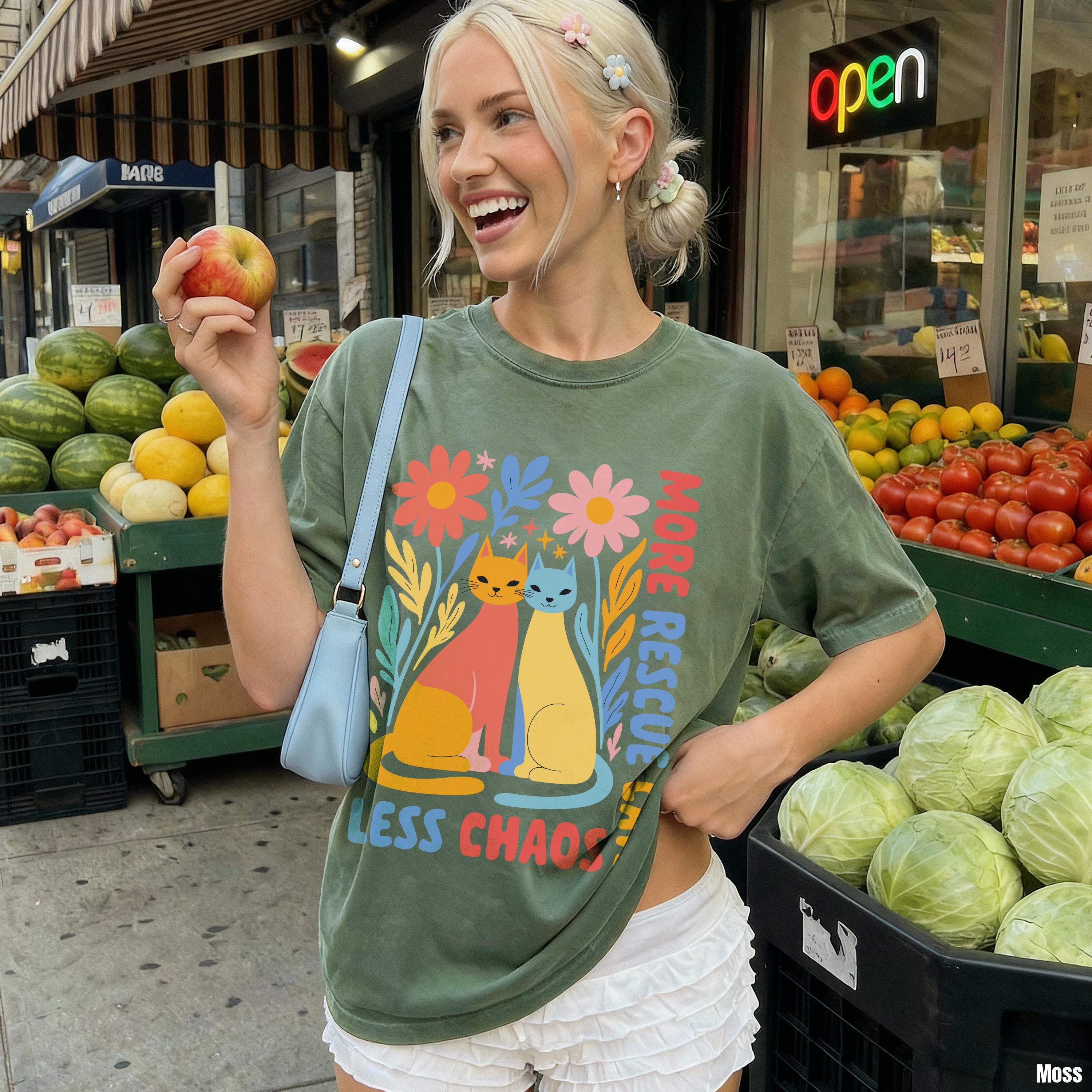 Woman in graphic t-shirt holding an apple, outdoor market produce in background.
