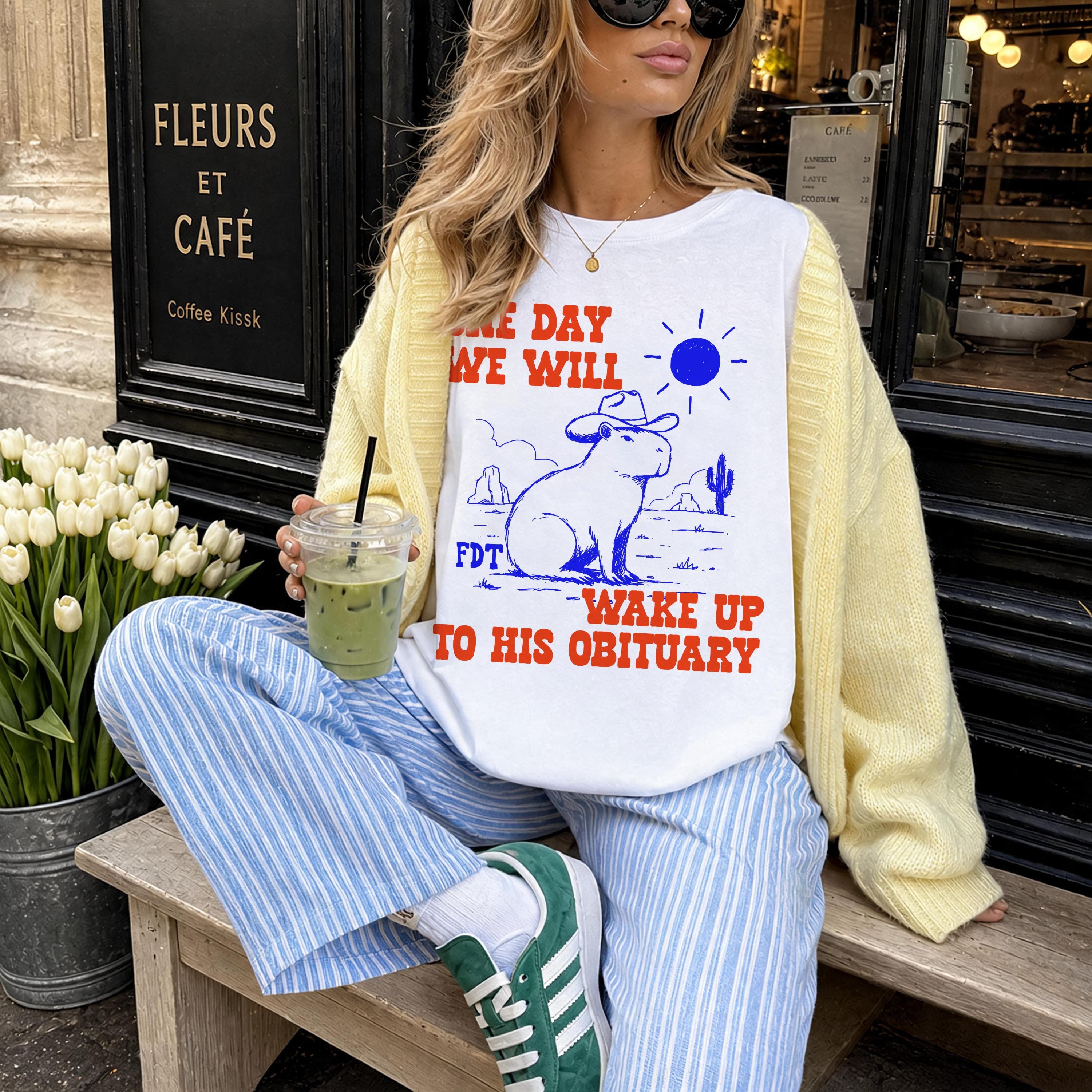White t-shirt with capybara graphic and text &amp;quot;ONE DAY WE WILL WAKE UP TO HIS OBITUARY&amp;quot;.