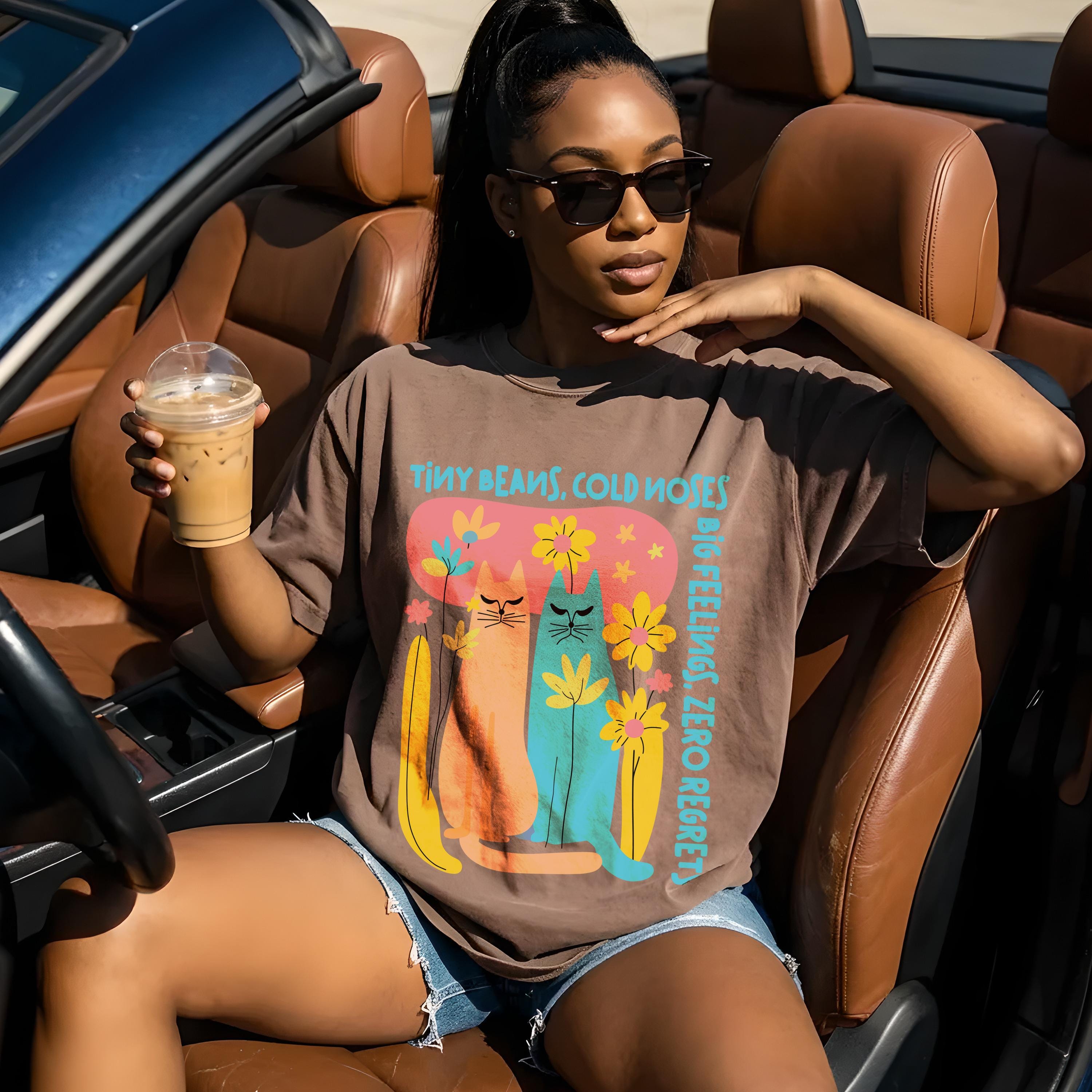 Woman in brown t-shirt with cats and text, holding iced coffee in car.