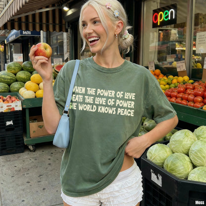 Woman in green t-shirt with text, holding an apple at a fruit stand with produce.