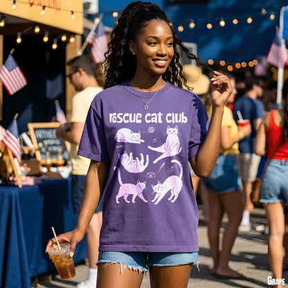 Woman in purple t-shirt with rescue cat illustrations and text, outdoors at an event.