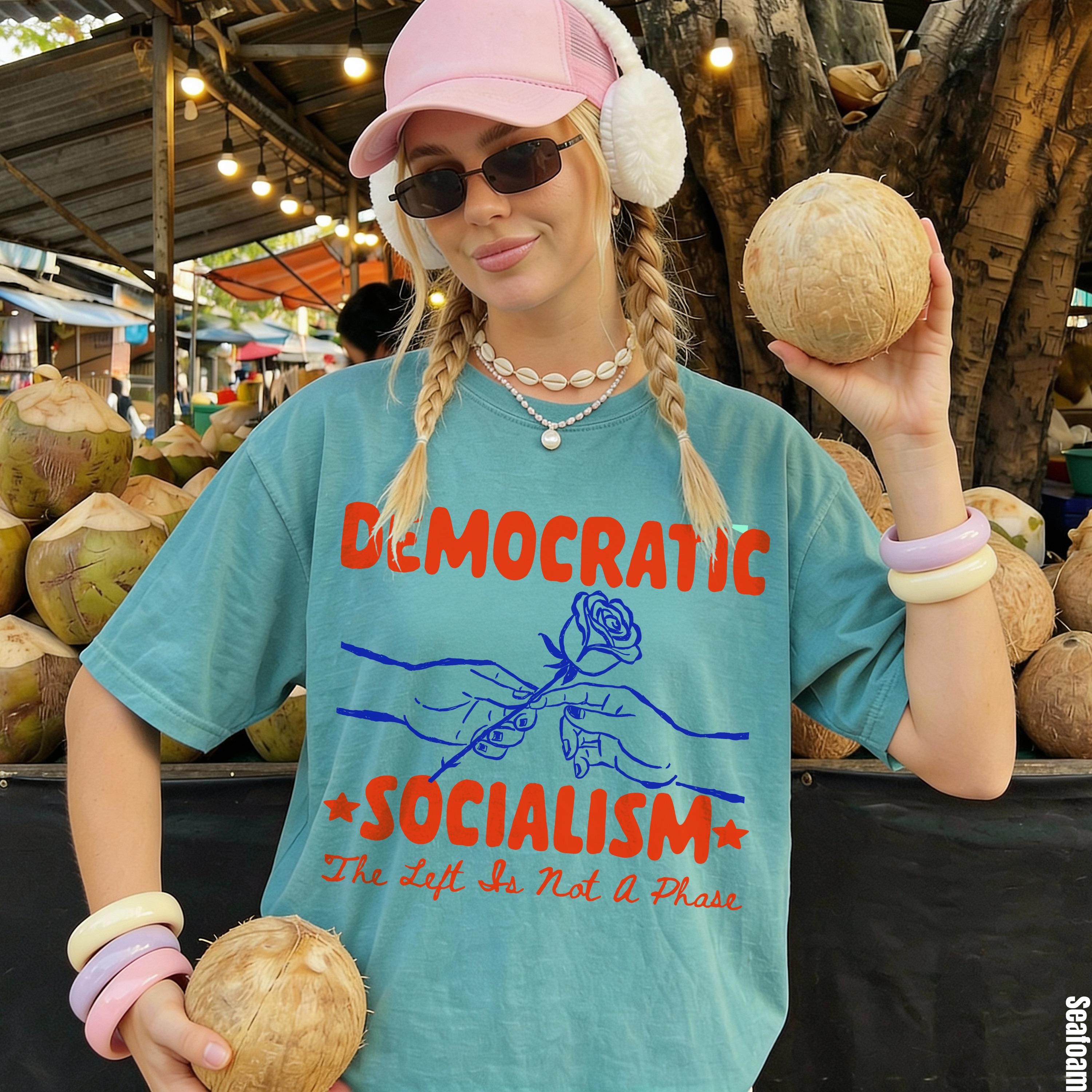 Teal t-shirt with &amp;quot;Democratic Socialism&amp;quot; and rose graphic, holding coconuts at market.