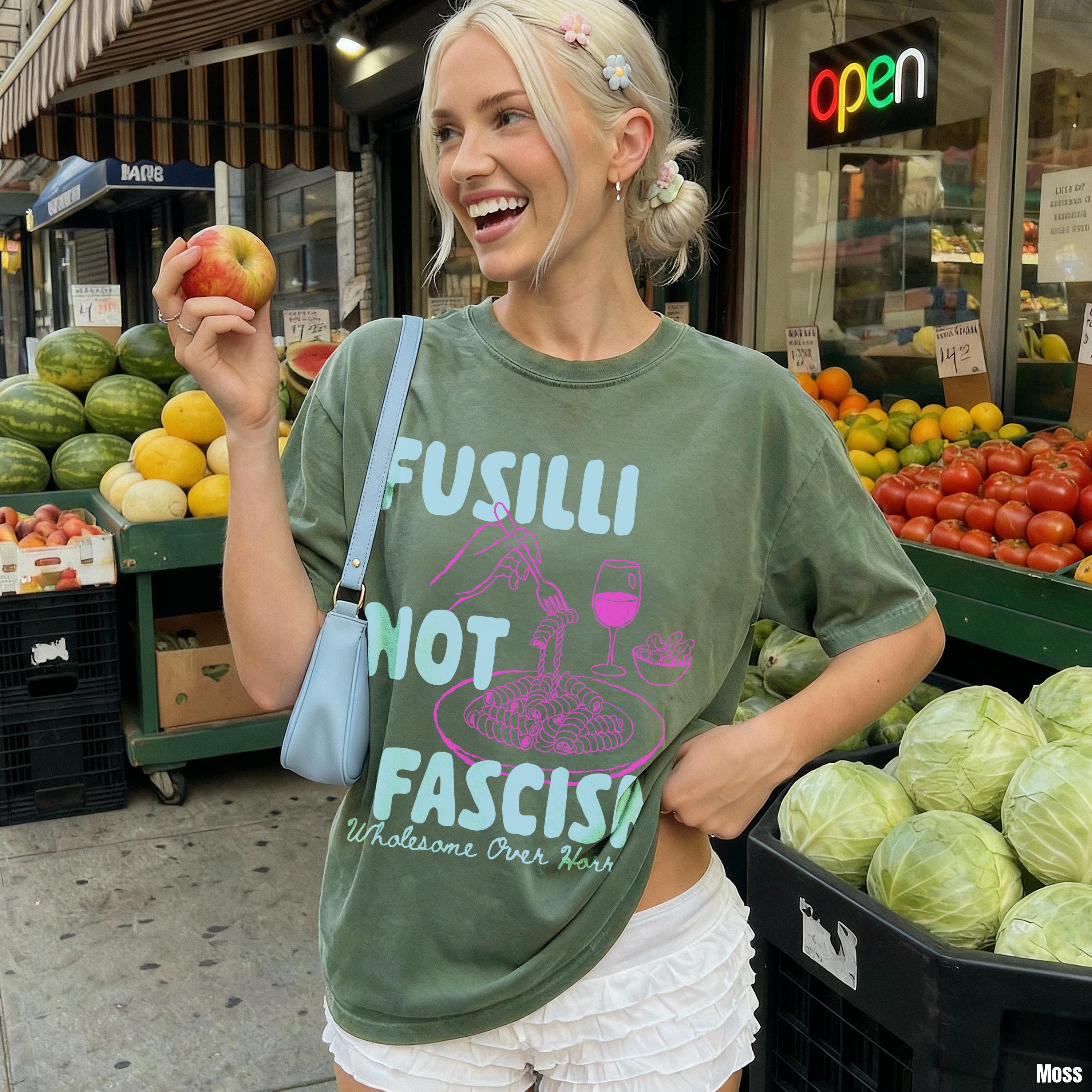 Woman in olive green shirt with &amp;quot;Fusilli Not Fascist&amp;quot; graphic holds an apple in front of a produce stand.