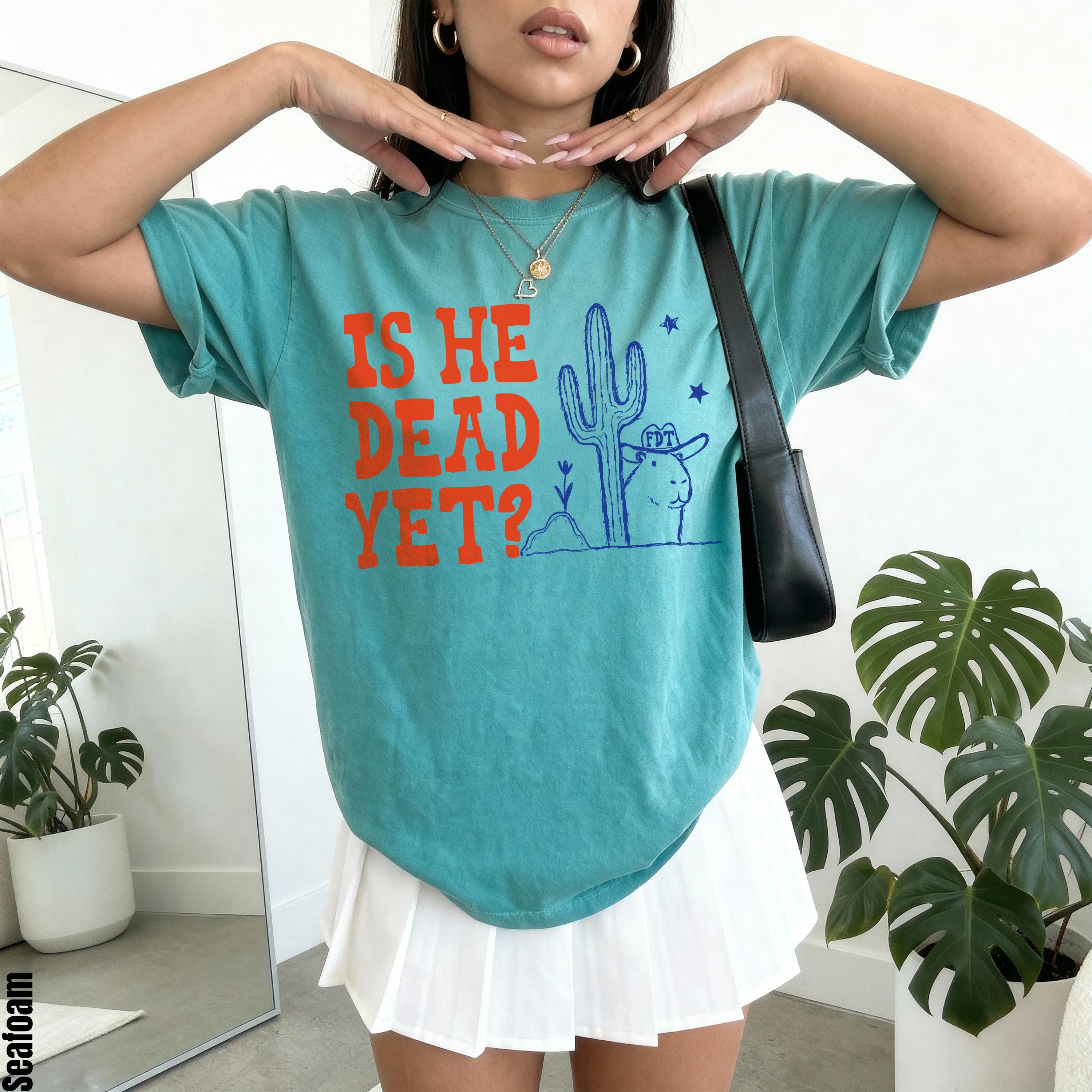 Teal t-shirt with orange text &amp;quot;IS HE DEAD YET?&amp;quot; and a cartoon desert design.