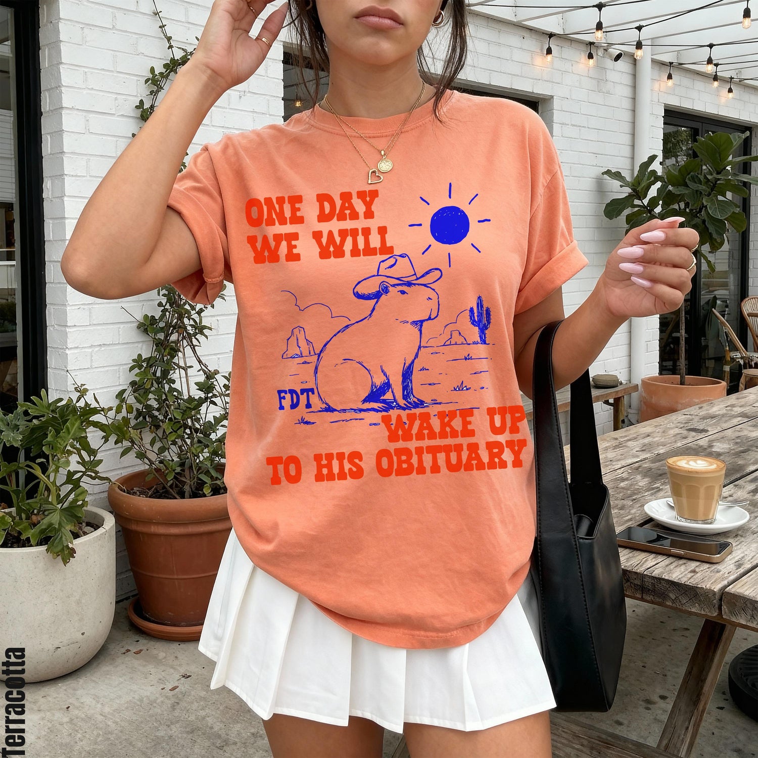 Orange t-shirt with blue graphic of capybara, cactus, and text &amp;quot;ONE DAY WE WILL WAKE UP TO HIS OBITUARY&amp;quot;.