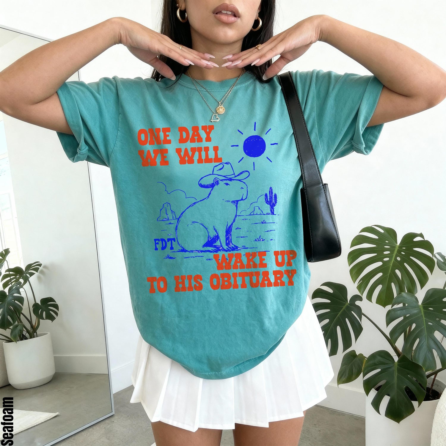 Teal t-shirt with &amp;quot;ONE DAY WE WILL WAKE UP TO HIS OBITUARY&amp;quot; and capybara graphic.