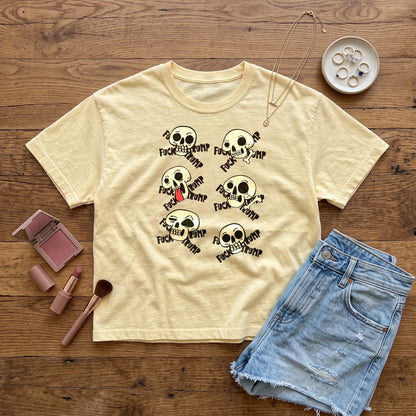 Light yellow t-shirt with multiple skull graphics saying &amp;quot;FUCK TRUMP&amp;quot; displayed on a wooden surface.