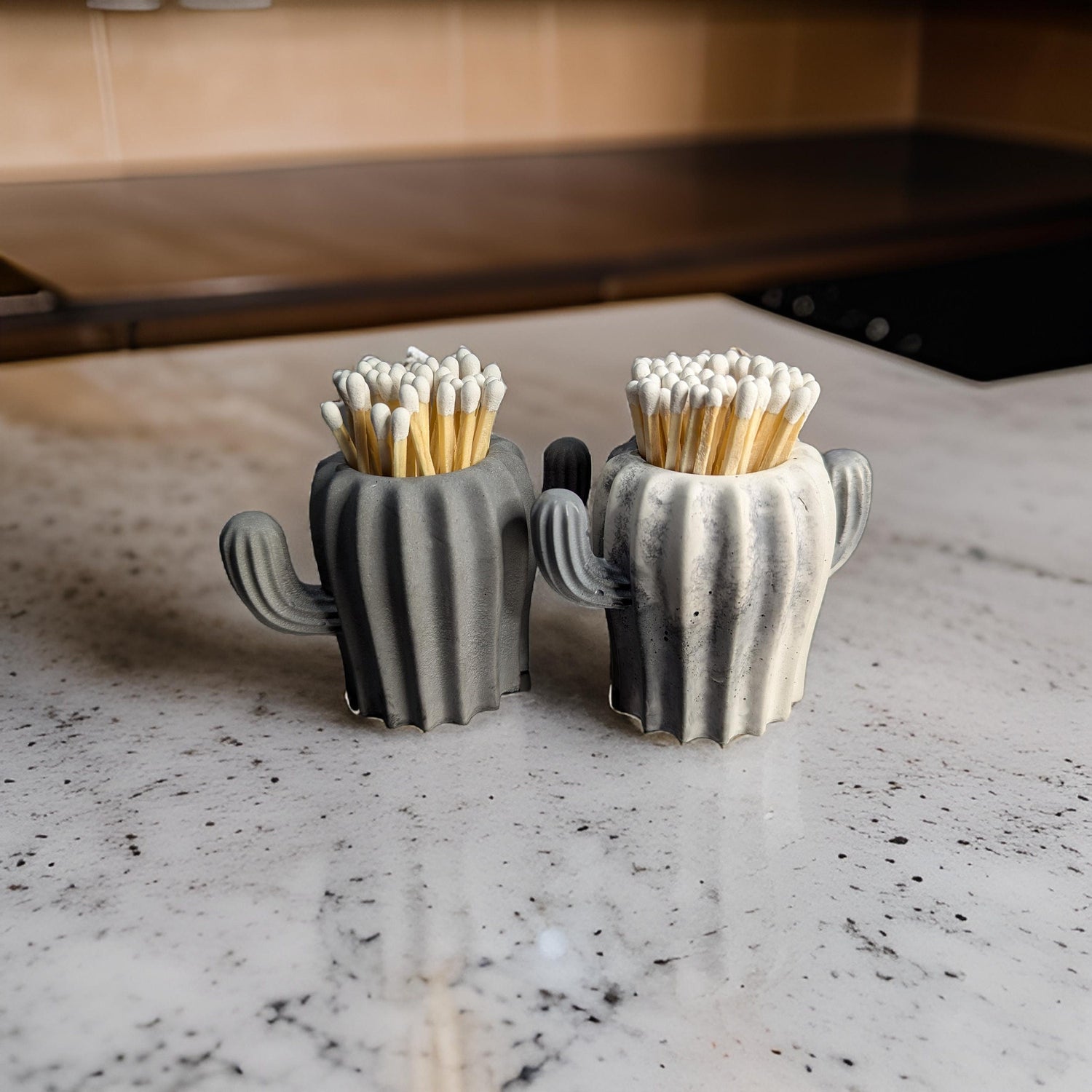 May include: Two gray ceramic cactus-shaped matchstick holders with white tips on the matches. The holders are on a white countertop.