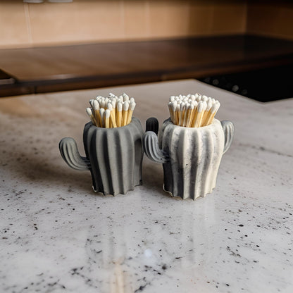 May include: Two gray ceramic cactus-shaped matchstick holders with white tips on the matches. The holders are on a white countertop.