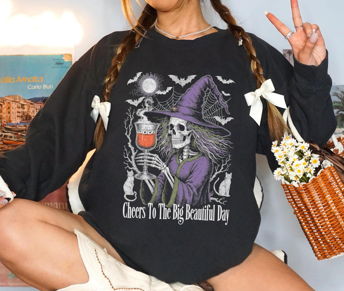 Black t-shirt with skeleton witch graphic and text &amp;quot;Cheers To The Big Beautiful Day&amp;quot;.