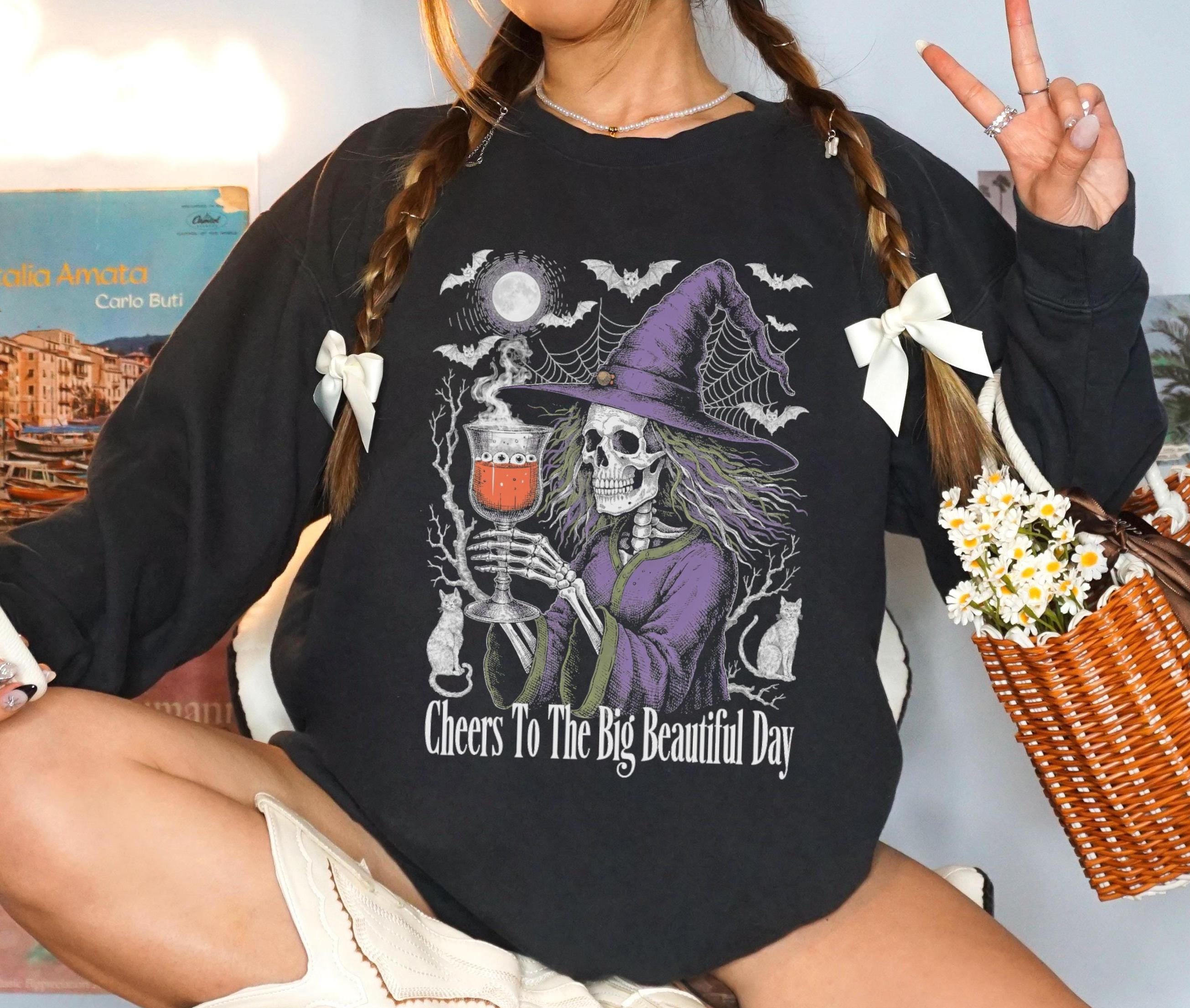 Black t-shirt with skeleton witch graphic and text &amp;quot;Cheers To The Big Beautiful Day&amp;quot;.