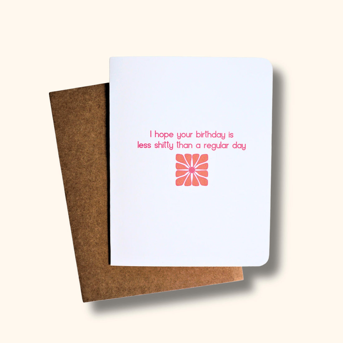 Funny white greeting card with “hope your birthday is less shitty than a regular day” quote in pink and orange font over a retro flower.