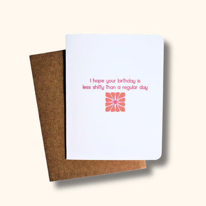 Funny white greeting card with “hope your birthday is less shitty than a regular day” quote in pink and orange font over a retro flower.