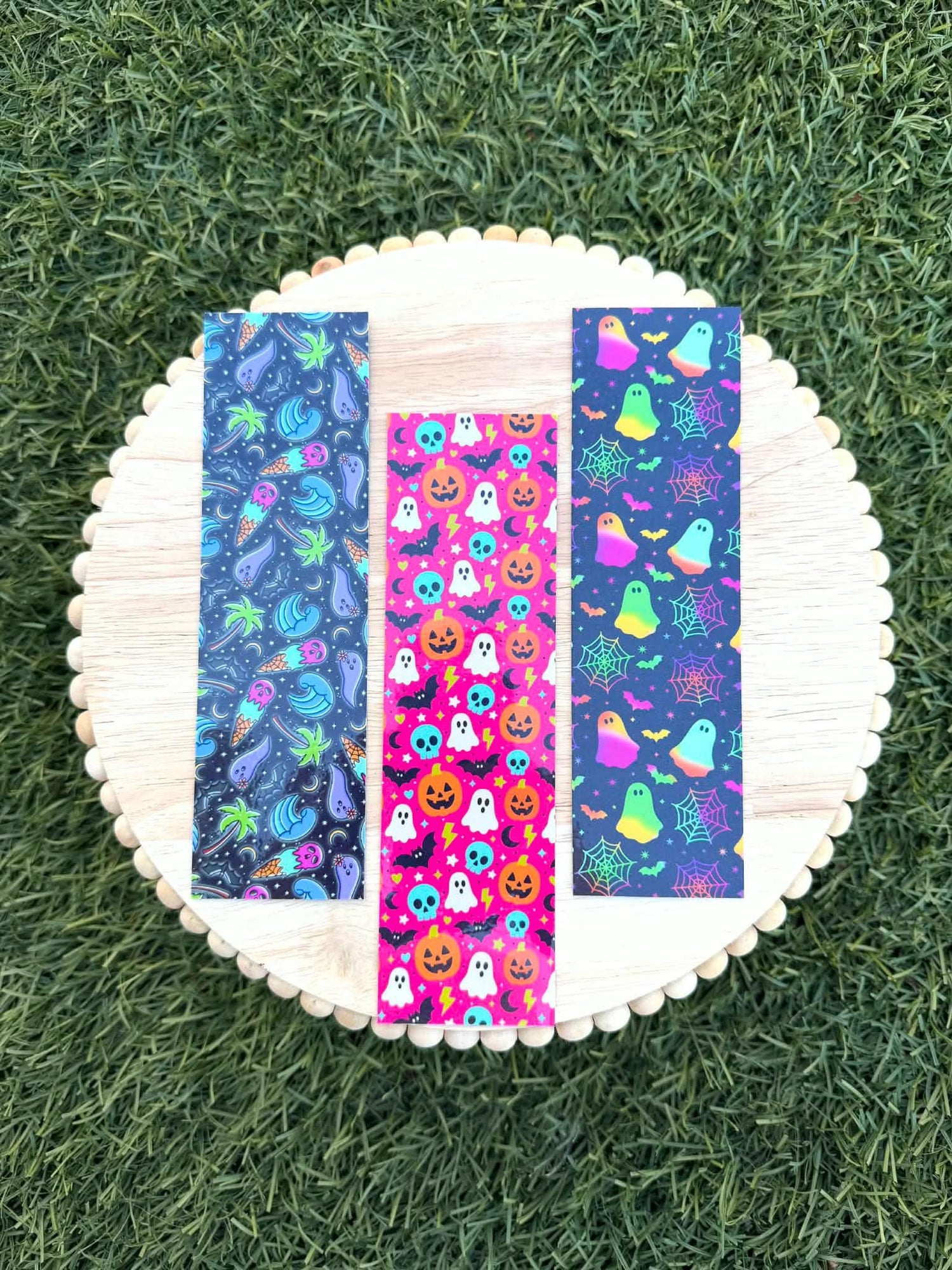 May include: Three rectangular bookmarks featuring vibrant patterns. The left bookmark has a dark background with neon palm trees and ice cream cones. The center bookmark has a bright pink background with orange pumpkins, white ghosts, and blue skulls. The right bookmark has a dark background with neon ghosts, spiderwebs, and bats.