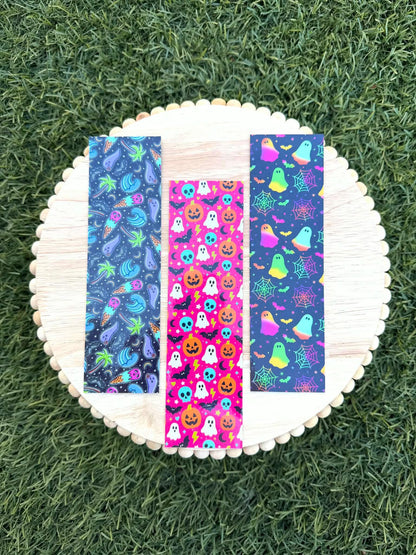 May include: Three rectangular bookmarks featuring vibrant patterns. The left bookmark has a dark background with neon palm trees and ice cream cones. The center bookmark has a bright pink background with orange pumpkins, white ghosts, and blue skulls. The right bookmark has a dark background with neon ghosts, spiderwebs, and bats.