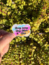 May include: A colorful sticker with the text "Busy doing Dog Mom Shit" in a playful font. The sticker features a paw print, a flower, and a smiling sun. The background is a vibrant green plant.