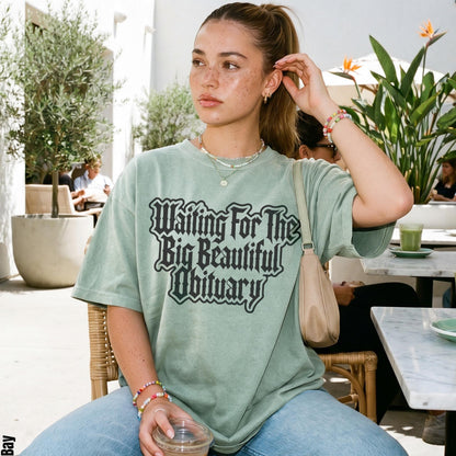 Woman wearing a green t-shirt with black gothic text that reads &amp;quot;Waiting For The Big Beautiful Obituary&amp;quot;.