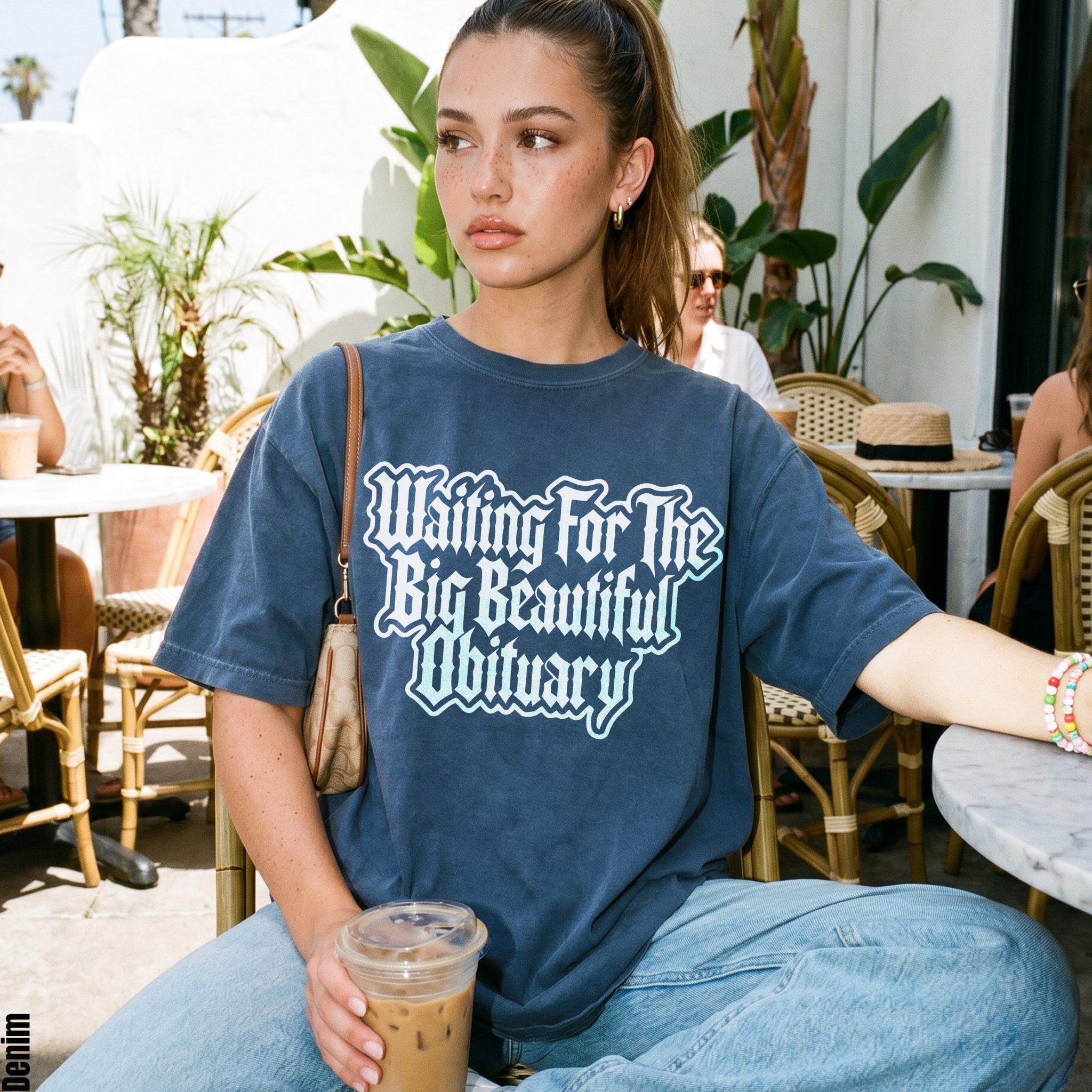 Woman wearing a dark blue t-shirt with &amp;quot;Waiting For The Big Beautiful Obituary&amp;quot; text.