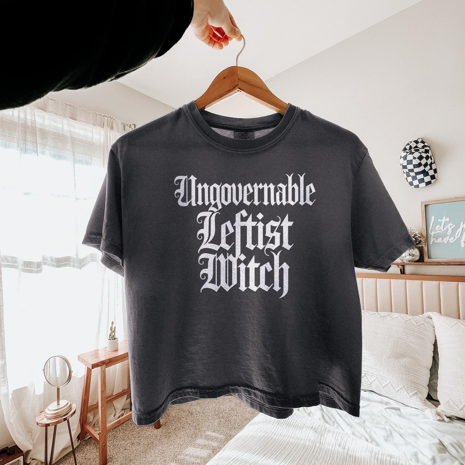 Dark grey t-shirt with white text &amp;quot;Ungovernable Leftist Witch&amp;quot; on a hanger.