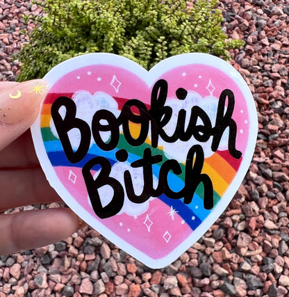 May include: A heart-shaped sticker with the words &amp;quot;Bookish Bitch&amp;quot; in black script. The sticker features a rainbow, clouds, and stars against a pink background. The sticker is held up by a hand.
