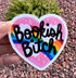 May include: A heart-shaped sticker with the words "Bookish Bitch" in black script. The sticker features a rainbow, clouds, and stars against a pink background. The sticker is held up by a hand.