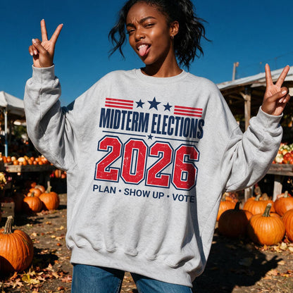 A woman in a grey crewneck sweatshirt featuring red and blue text reading MIDTERM ELECTIONS 2026 PLAN SHOW UP VOTE poses with peace signs at a pumpkin patch.