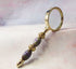 The Color Changing Magnifying Glass: Boho Office Decor features an ornate gold and purple handle with intricate patterns—ideal for mindful study and adding boho flair to your workspace. Shown resting on a soft, pastel-colored surface.
