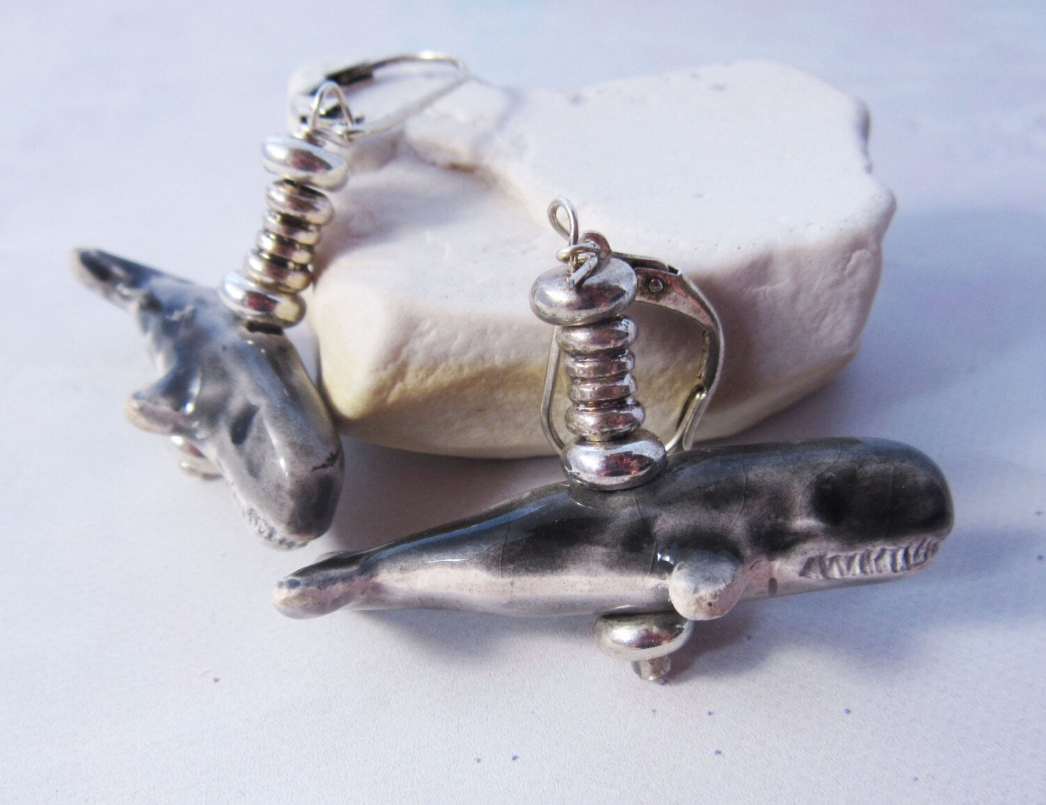 The Hand-Painted Sperm Whale Earrings—sterling silver ocean jewelry—showcase artisan whale motifs with metallic bead accents, displayed on a white stone.