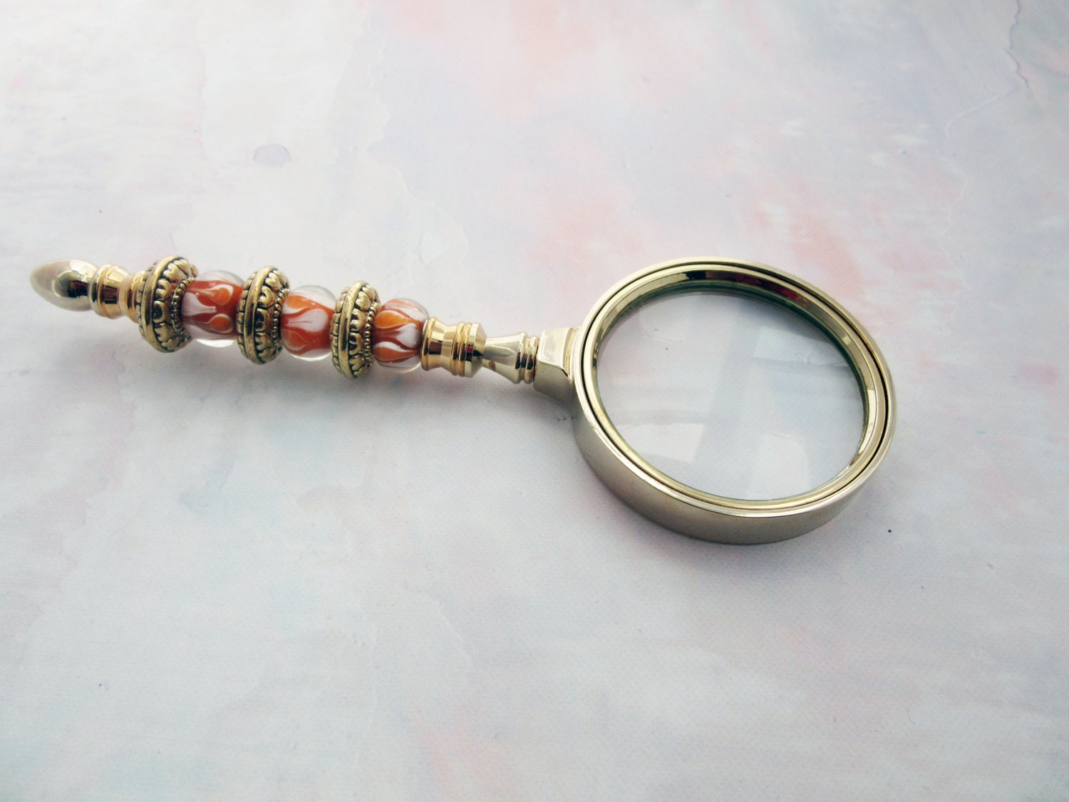 The Orange and White Glass Magnifier features a beaded handle with artistic details, making it an ideal gift for art lovers. Its elegant design stands out on any pastel-hued surface.