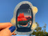 a hand with a painted nail holds a Sedona Sunset Lantern up to the sky in the sunset light. The stickers shows a sunset over the red rocks of Sedona with trees and brush next to a stream. There are mountains in the distance and a crescent moon in the sky. This scene is inside a lantern. 