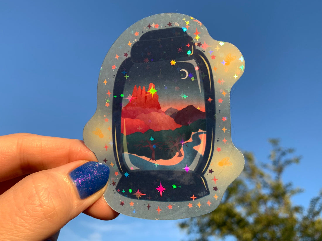 a hand with a painted nail holds a Sedona Sunset Lantern up to the sky in the sunset light. The stickers shows a sunset over the red rocks of Sedona with trees and brush next to a stream. There are mountains in the distance and a crescent moon in the sky. This scene is inside a lantern. 