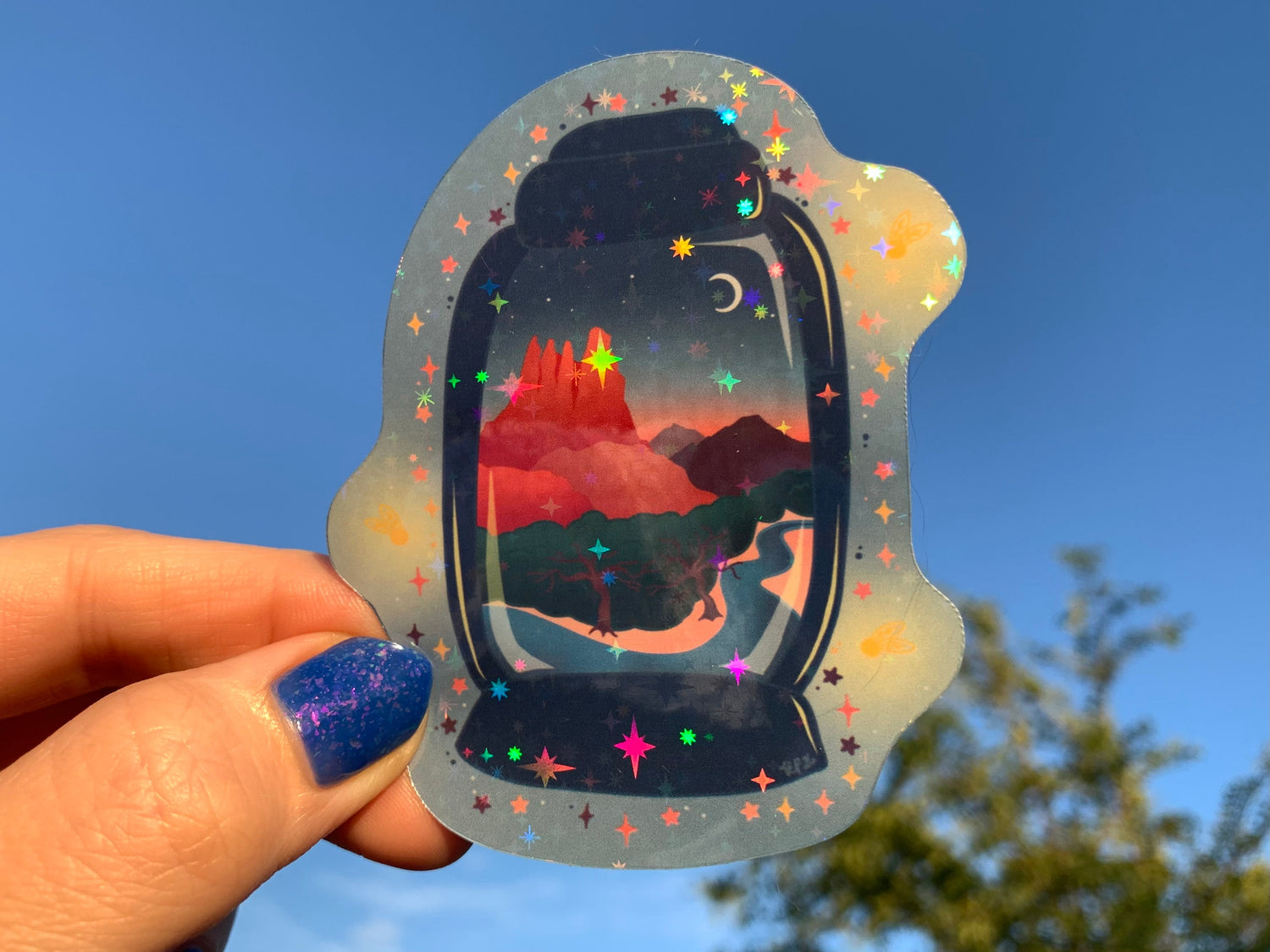 a hand with a painted nail holds a Sedona Sunset Lantern up to the sky in the sunset light. The stickers shows a sunset over the red rocks of Sedona with trees and brush next to a stream. There are mountains in the distance and a crescent moon in the sky. This scene is inside a lantern. 