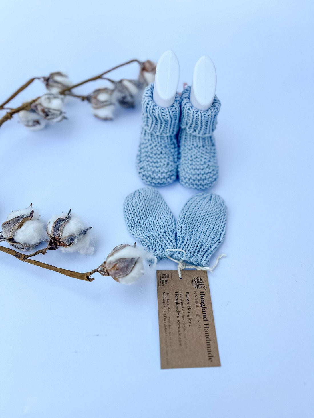 A pair of light blue, hand-knit organic cotton baby booties and mittens are displayed in a baby foot mold on a white background with a cotton stem on its left in the foreground and background