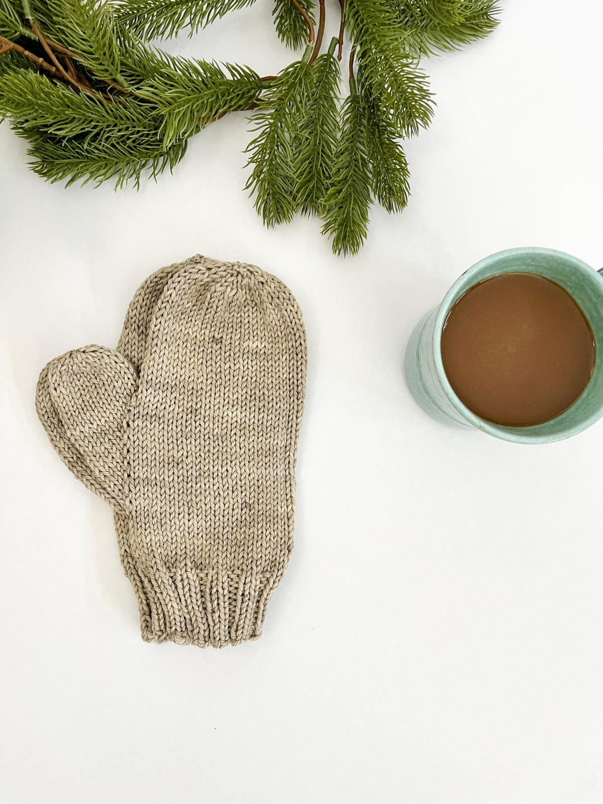 A pair of gray colored, hand-knit, hand-dyed merino dyed with plants, is displayed on a white background with a cup of coffee and some evergreen greenery in the background