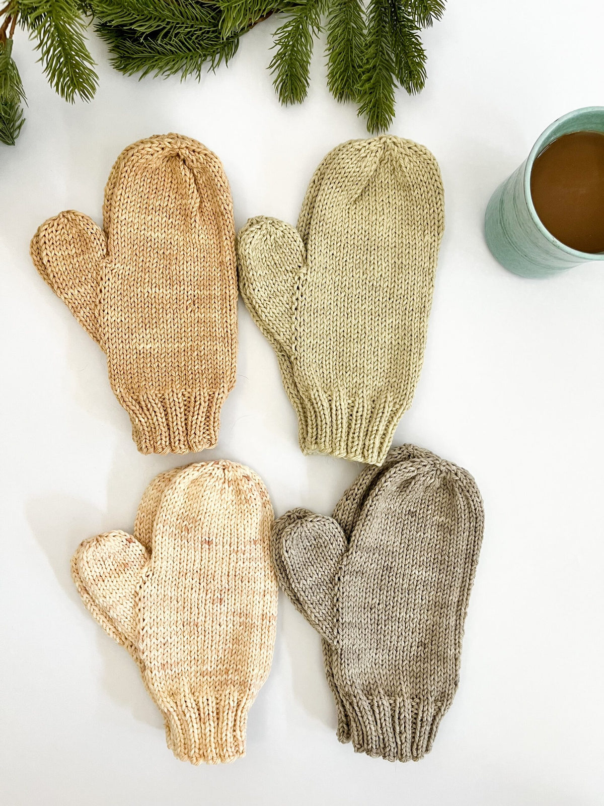 Four pairs of hand-knit, hand-dyed merino dyed with plants, are displayed on a white background with a cup of coffee and some evergreen greenery in the background. The mittens are tan, eucalyptus, cream, and gray.