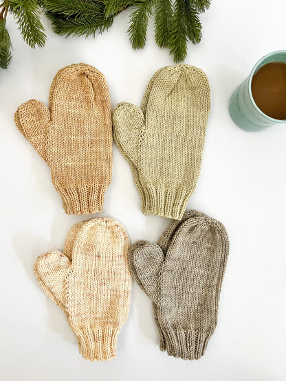 Four pairs of hand-knit, hand-dyed merino dyed with plants, are displayed on a white background with a cup of coffee and some evergreen greenery in the background. The mittens are tan, eucalyptus, cream, and gray.