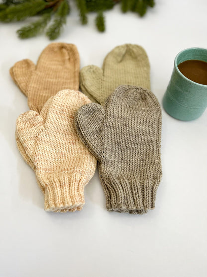 Four pairs of hand-knit, hand-dyed merino dyed with plants, are displayed on a white background with a cup of coffee and some evergreen greenery in the background. The mittens are tan, eucalyptus, cream, and gray.