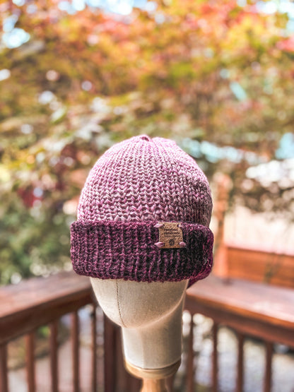 A hand-knit, double-layer beanie with a darker mauve brim and lighter colored mauve head is displayed on a wood and cloth mannequin head in the foreground of the photo. In the background, there is a wood railing and a maple tree with fall colors