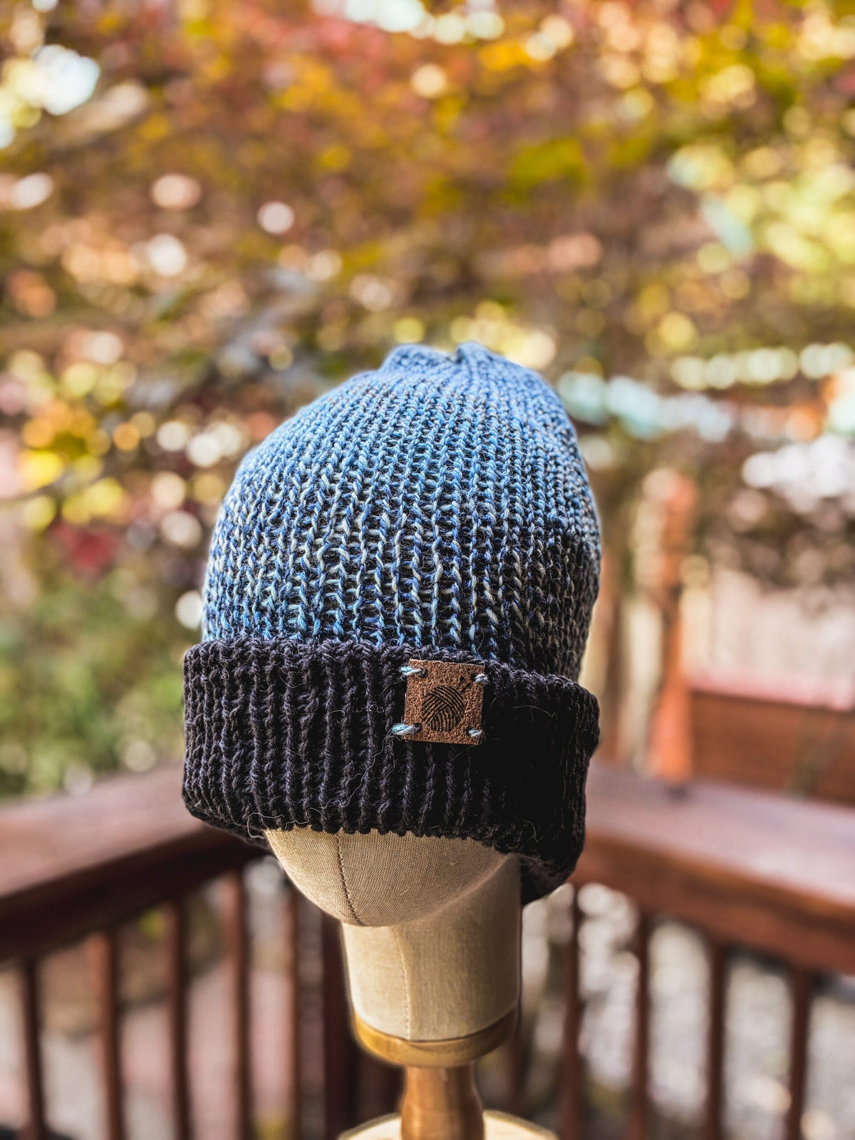 A hand-knit, double-layer beanie with a darker blue brim and lighter blue and purple ombre head is displayed on a wood and cloth mannequin head in the foreground of the photo. In the background, there is a wood railing and a maple tree with fall colors