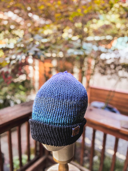A hand-knit, double-layer beanie with a darker blue brim and lighter blue and purple ombre head is displayed on a wood and cloth mannequin head in the foreground of the photo. In the background, there is a wood railing and a maple tree with fall colors