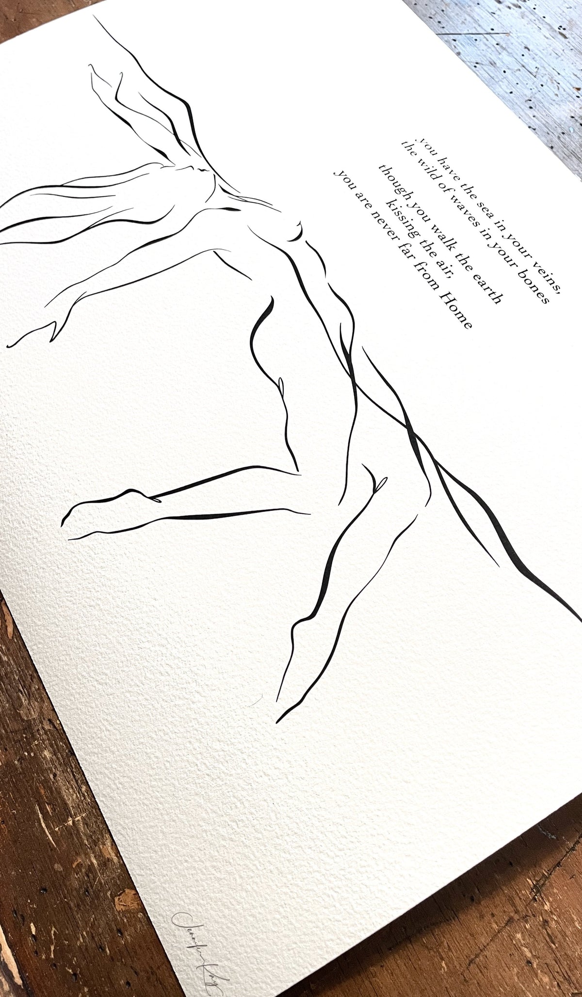 Black and white elegant line art illustration of sensual woman floating in water; fine art print on slightly textured Aquarelle Rag paper lays on wooden desk.