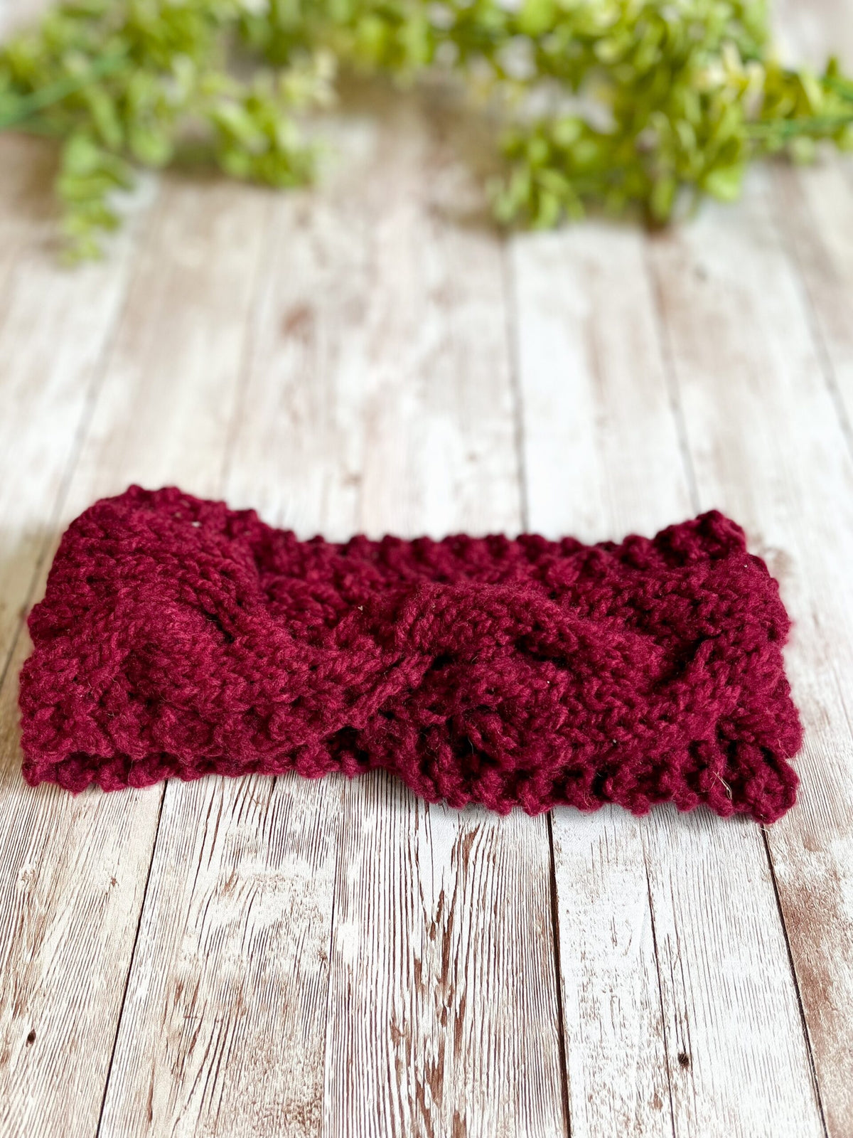 A red, Virginia-raised merino ear warmer rests on a wood plank with greenery in the background