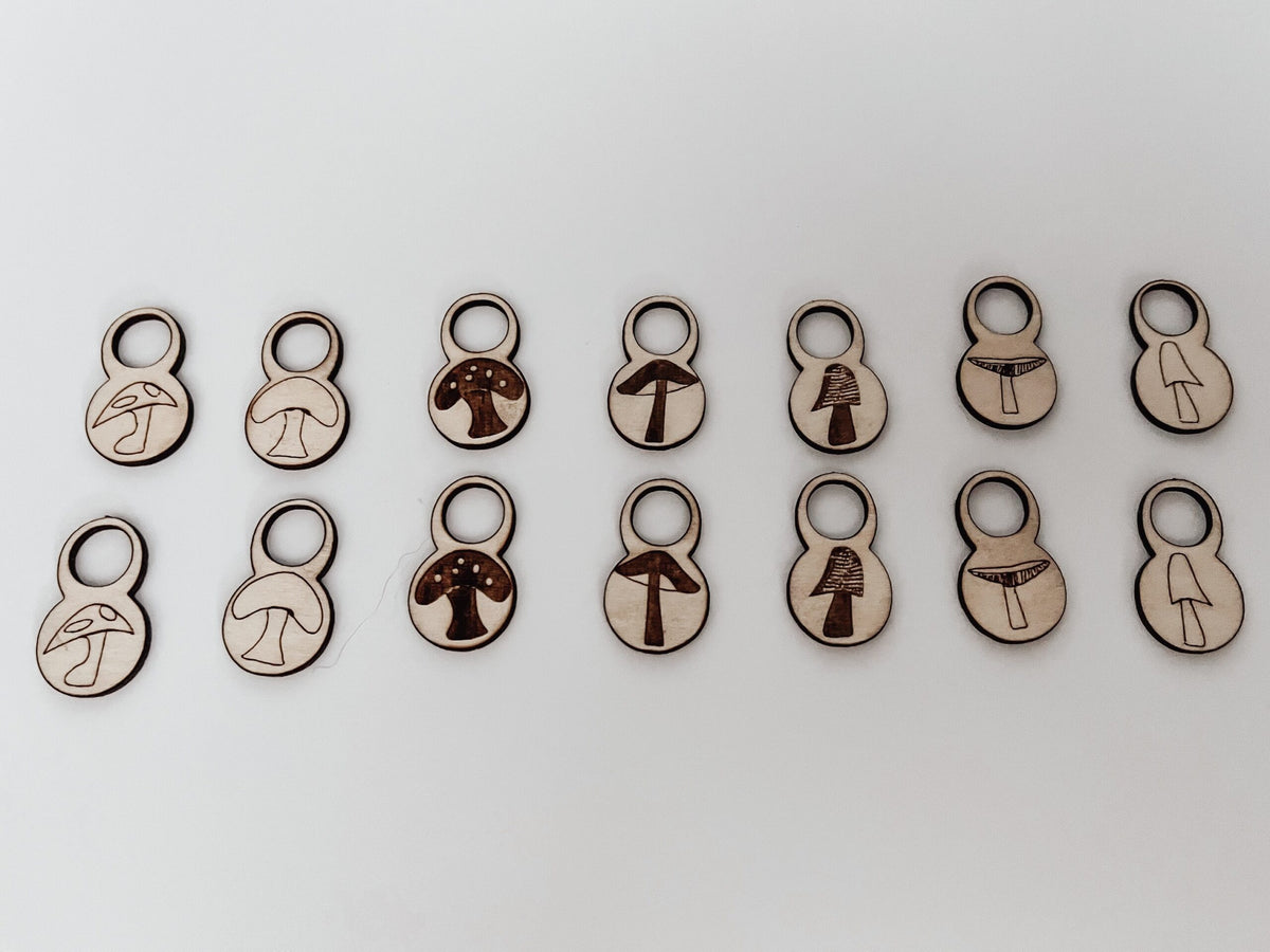 A total of 14 wood stitch markers with a variety of hand-drawn mushroom designs are displayed on a white background.