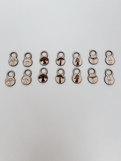 A total of 14 wood stitch markers with a variety of hand-drawn mushroom designs are displayed on a white background.