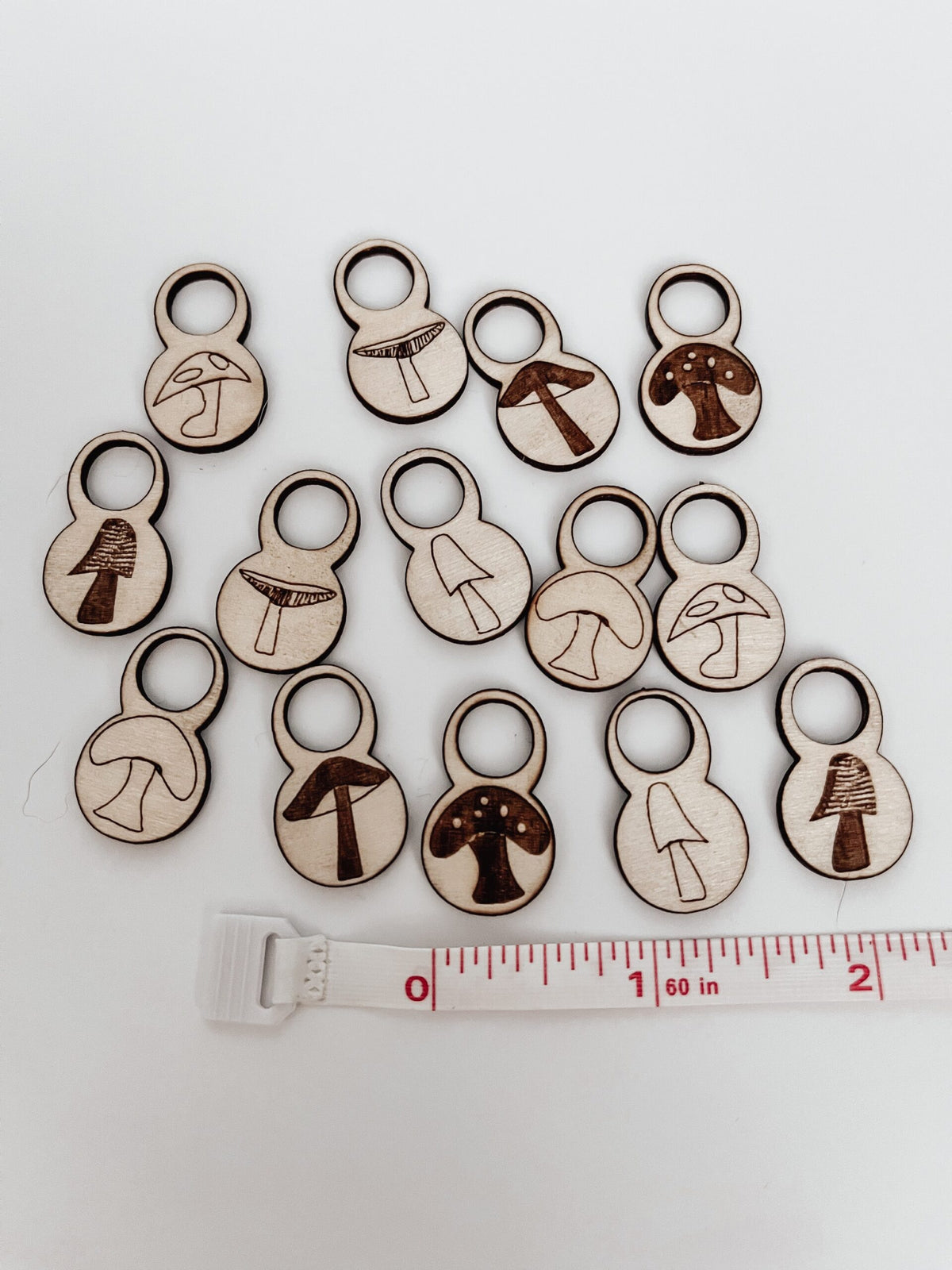 A total of 14 wood stitch markers with a variety of hand-drawn mushroom designs are displayed on a white background. next to a measuring tape showing they&